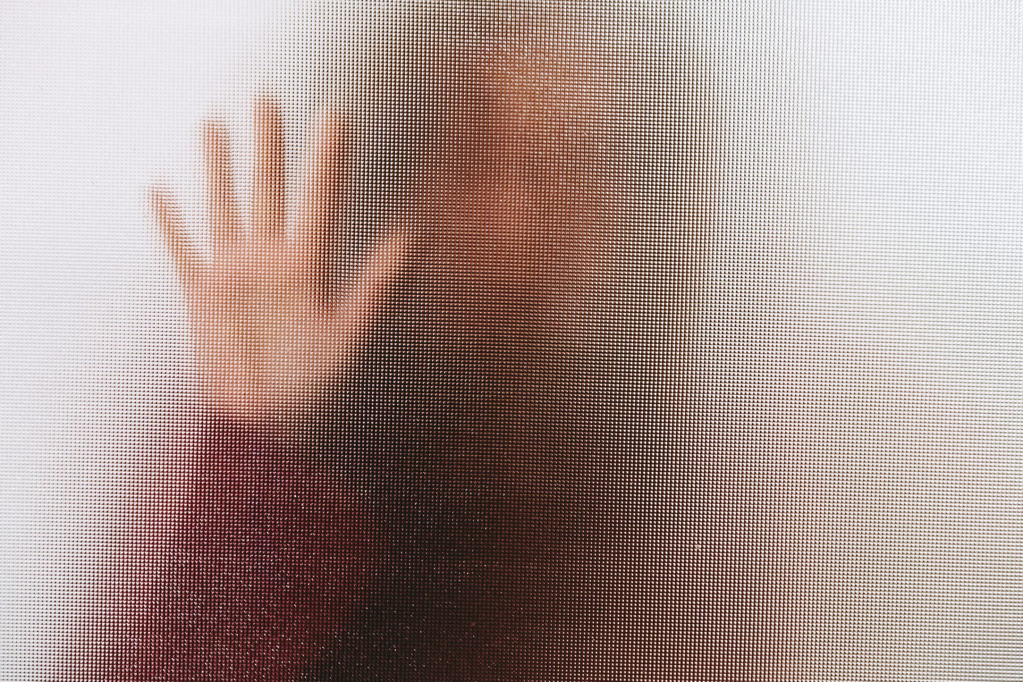 Back lit image of the silhouette of a woman with her hands pressed against a glass window. The silhouette is distorted, and the arms elongated, giving an alien-like quality.