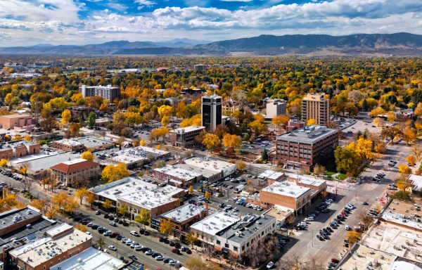 Aerial of Downtown Fort Collins during autumn, with downtown buildings and the lush city green and golden urban treescape in the foreground, and the Rocky Mountains and a cloudscape in the background.