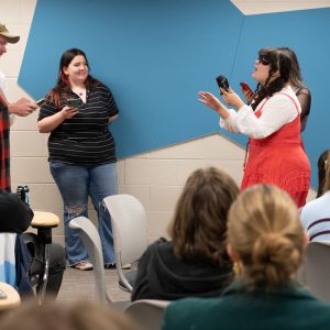 Students perform scenes in front of the classroom, reading lines off their mobile phones