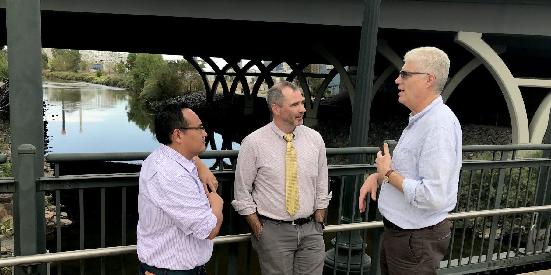 Chris Conner is standing with Eric Aoki and Greg Dickinson on a pedestrian bridge that crosses the South Platte River near Colfax Avenue