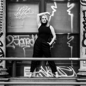 black and white image of a women in a black jumper posing with one arm over her head in front of a building covered in graffiti